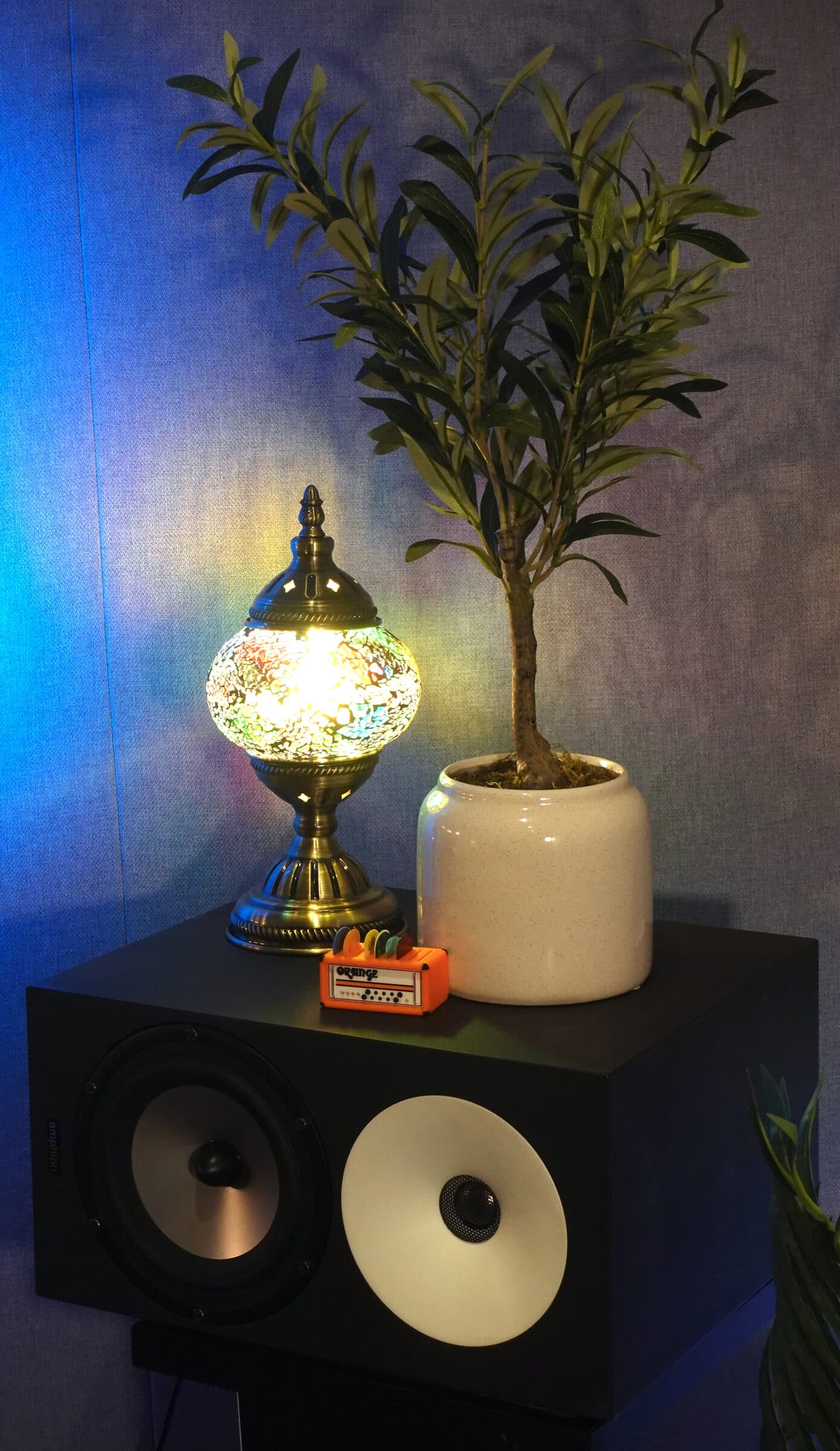 A monitor cabinet in the corner of the room beneath a mosaic lamp and olive tree